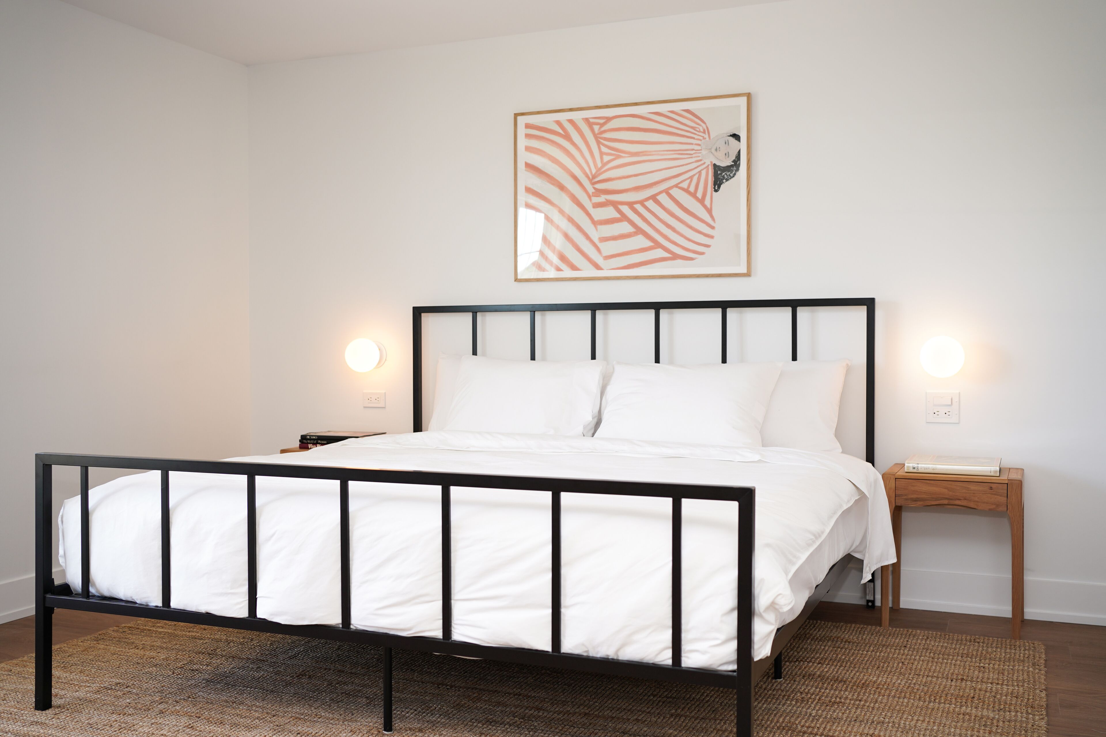 Una cama bien tendida con estructura de metal negro y ropa de cama blanca, una mesita de noche de madera con una lámpara, y una obra enmarcada en la pared.