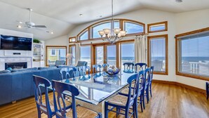 A modern kitchen with a large island, a dining area with blue chairs, and a view of the ocean through large windows. - Sand Pebbles: Oceanfront, Pool, Hot Tub, Rec Room (Nags Head)