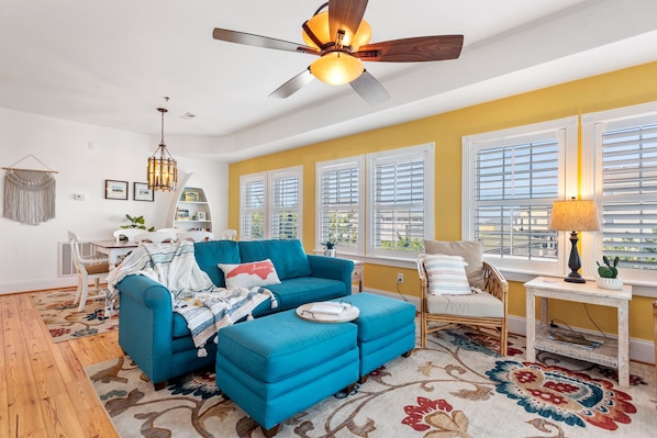 A bright living room with a blue sofa, a ceiling fan, and large windows with blinds.