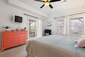 A bedroom with a bed, a red dresser, a ceiling fan, a TV, and a window with blinds.