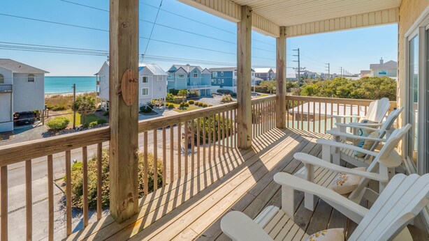 Eine Holzterrasse mit weißen Stühlen, mit Blick auf einen Strand und Wohnhäuser.