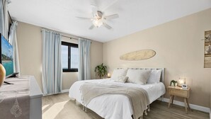 A bedroom with a bed, a bedside table, a ceiling fan, a window with curtains, and a wall-mounted artwork.