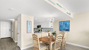A dining area with a wooden table and chairs, a chandelier, and a kitchen in the background.