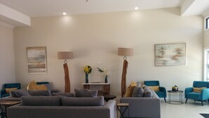 Un salon moderne avec des canapés gris, des fauteuils bleus et une table de ping-pong. On y trouve deux lampadaires, un vase avec des fleurs et un tableau accroché au mur.