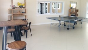 Une pièce spacieuse équipée d’une table de ping-pong, d’une table à manger avec des chaises et d’une grande fenêtre avec des stores.