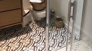 A bathroom with a white sink, a toilet, a wicker basket on the toilet, a glass shower enclosure, and patterned floor tiles. - Charming single-storey studio in a quiet location  (La Chapelle-Saint-Mesmin)