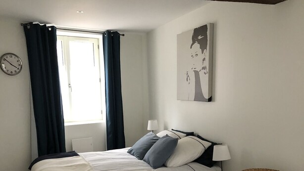 A bedroom with a bed, a chair, a table, a clock, and a window with curtains. - Charming single-storey studio in a quiet location  (La Chapelle-Saint-Mesmin)