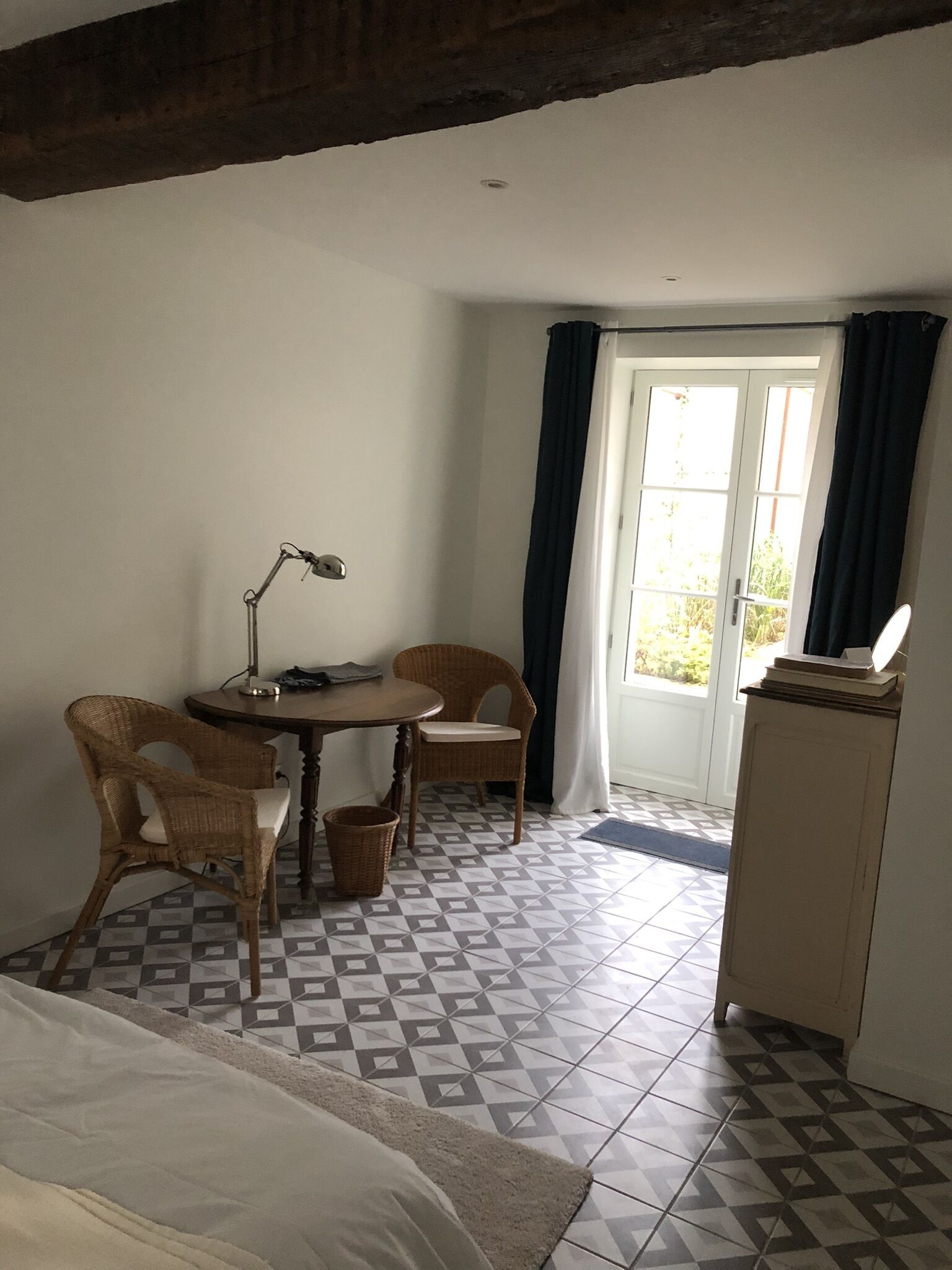 Una habitación con una cama, un escritorio con lámpara, sillas de mimbre, un armario y un suelo estampado.