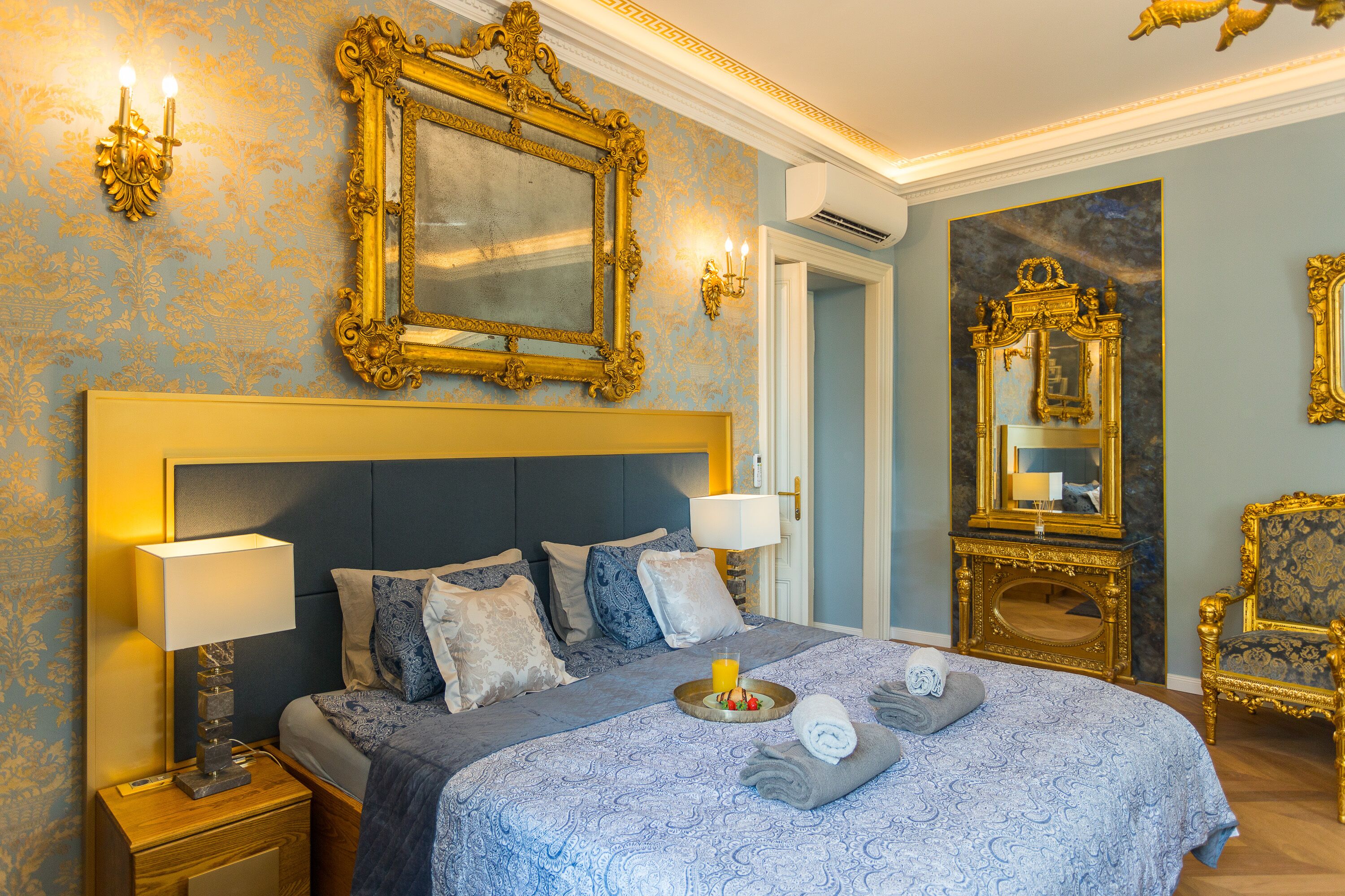 Une chambre d’hôtel avec un grand lit, deux lampes de chevet, un plateau avec des serviettes et un verre de jus d’orange, ainsi que des miroirs décoratifs encadrés d’or.