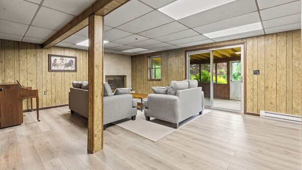A spacious room with wooden walls, a piano, two grey sofas, a wooden coffee table, and a large sliding glass door leading to an outdoor area.