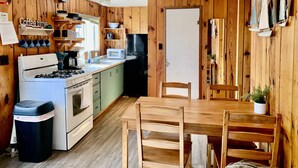 Kompakti keittiö, jonka seinät ovat puuta, ja jossa on liesi, mikroaaltouuni, jääkaappi sekä ruokapöytä tuoleineen.