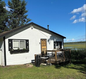 Valkoinen mökki, jossa on puinen terassi, ovi ja ikkunat, joissa on sälekaihtimet.