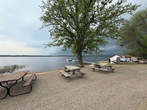 Järvenrantainen piknikalue, jossa on puisia penkkejä ja piknikpöytiä, suuri puu ja taustalla pieni rakennus.