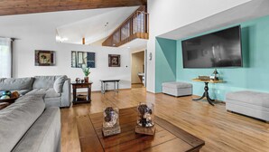 A spacious living room with a wooden ceiling, a grey sofa, a coffee table, and a TV mounted on a teal wall.