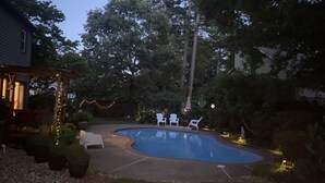 Un jardin à l’arrière de la maison, avec une piscine, entouré d’arbres et de plantes en pot. On y trouve des chaises d’extérieur et une pergola illuminée de guirlandes lumineuses.