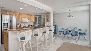 Une cuisine moderne avec des meubles blancs, un plan de travail en granit, des appareils en acier inoxydable et un coin repas comprenant une table en verre et des chaises bleues.