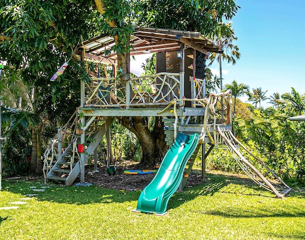 Un espace de jeux en forme de cabane dans les arbres, avec un toboggan, des structures d’escalade et une plateforme en bois.
