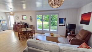 En rummelig opholdsstue med et træspisebord, en sofa, et fjernsyn og et stort vindue med udsigt til grøn natur.