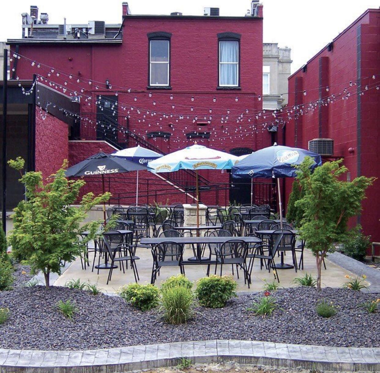 Une terrasse extérieure avec des tables et des parasols, entourée d’un jardin agrémenté de petits arbres et d’arbustes.