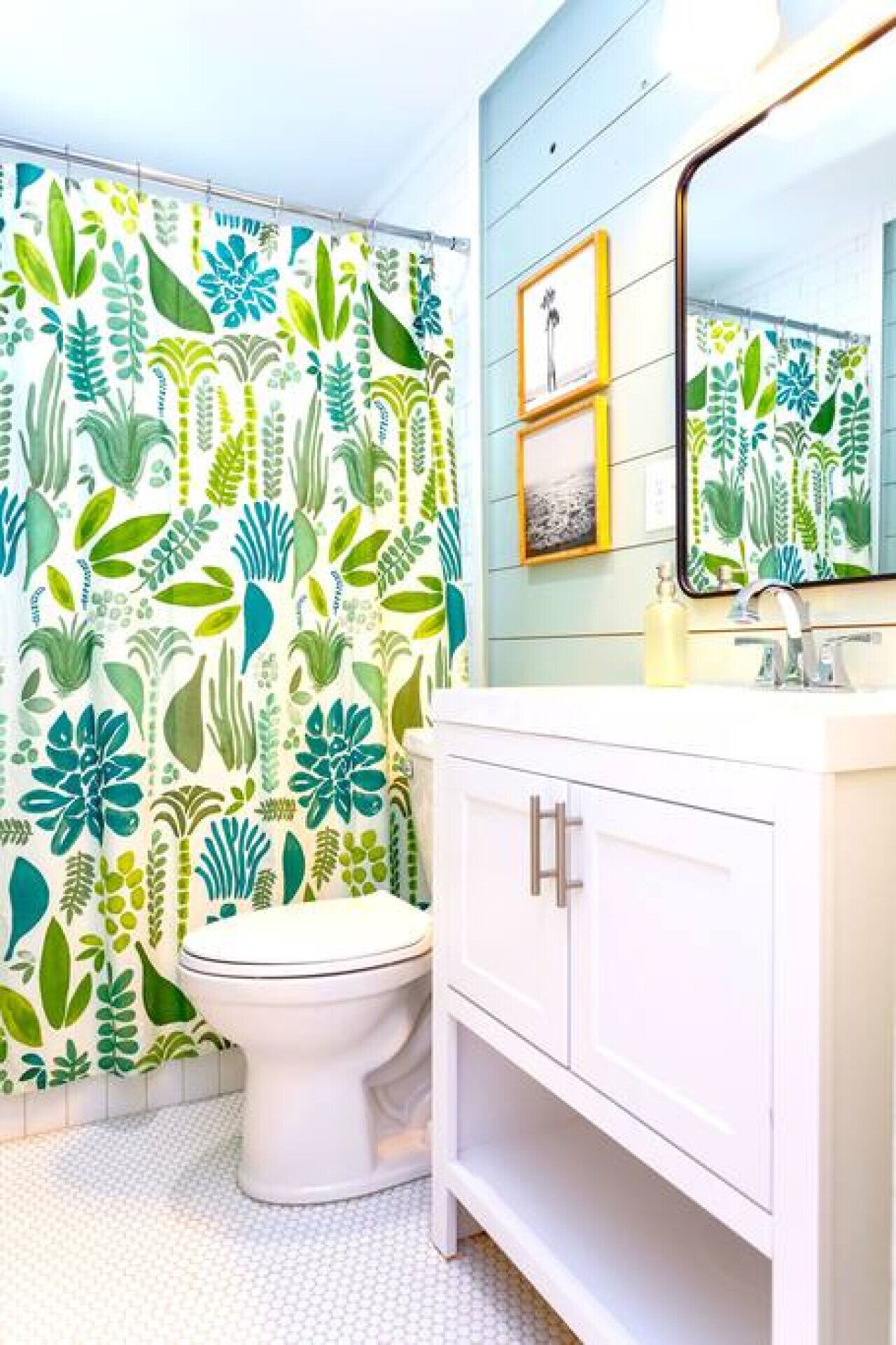 A bathroom with a toilet, a white vanity, a mirror, and a shower curtain with a leafy pattern.