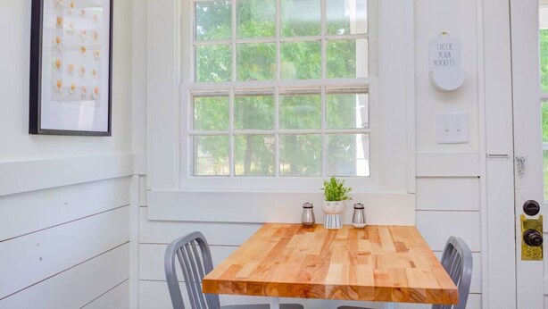 A dining area with a wooden table, two chairs, a framed picture, a window with a view of greenery, and a sign on the door. - Charming parkview from rocking chair front porch (Graham)