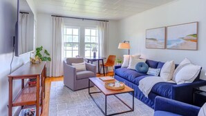 A living room with a blue sofa, grey armchair, and a small table with a tray. There are two windows with curtains, a lamp, and a TV mounted on the wall. - Charming parkview from rocking chair front porch (Graham)
