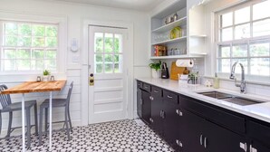 A kitchen with a black and white tiled floor, a wooden table with chairs, a white door, and a window with a view of greenery. - Charming parkview from rocking chair front porch (Graham)