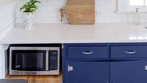 Una cocina con armarios blancos, una tabla de cortar de madera, un microondas y una estantería con utensilios de cocina y un cartel que dice '¡Hola!'.