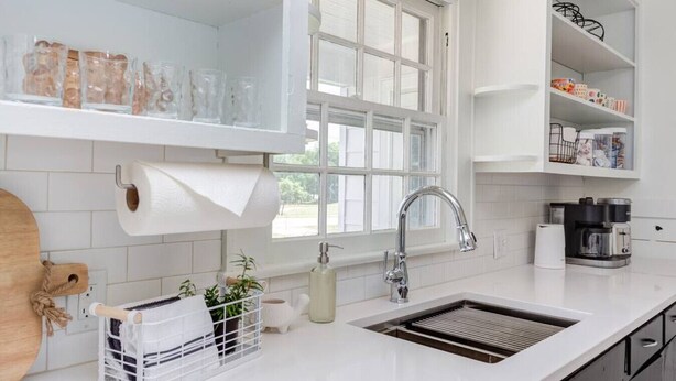 Una cocina moderna con armarios blancos, un ventanal grande, un lavamanos de acero inoxidable y una cafetera.