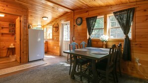 A wooden cabin interior with a dining table, chairs, a refrigerator, and a clock on the wall. - Pine Ridge Cabin: an all-seasons Wellsboro getaway (Charleston Township)