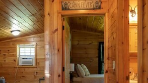 A cabin interior with a bed, a small table, a window with an air conditioner, and a sign reading "CABIN FEVER". - Pine Ridge Cabin: an all-seasons Wellsboro getaway (Charleston Township)