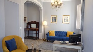 A living room with a yellow armchair, a blue sofa, a wooden desk, a chandelier, and a television.