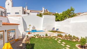 A white-walled building with a swimming pool, a small garden, and a paved area with lounge chairs.
