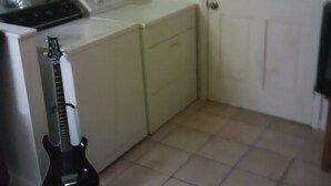 Une cuisine avec une porte blanche, un sol carrelé et une guitare noire appuyée contre le mur.