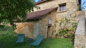 A stone building with a sloped roof, a small window, and a wooden shutter. There are two green chairs outside.