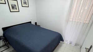 Een slaapkamer met een bed, nachtkastje, wandluidspreker en raam met dunne gordijnen.