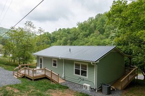 A small, single-story cabin with a metal roof, a wooden deck, and a gravel driveway.