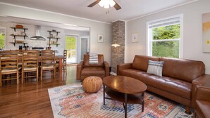 A spacious living room with a brown leather sofa, a wooden coffee table, and a patterned rug. A ceiling fan and a fireplace are visible in the background. - 4 BR ~ 4 Mins to Hatley Pointe ~ Fall Mtn Views (Mars Hill)