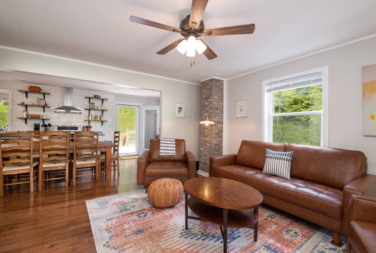 A spacious living room with a brown leather sofa, a wooden dining table with chairs, a ceiling fan, and a large window with a view of greenery.