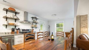 A modern kitchen with white cabinets, a wooden dining table, and a brick accent wall. - 4 BR ~ 4 Mins to Hatley Pointe ~ Fall Mtn Views (Mars Hill)