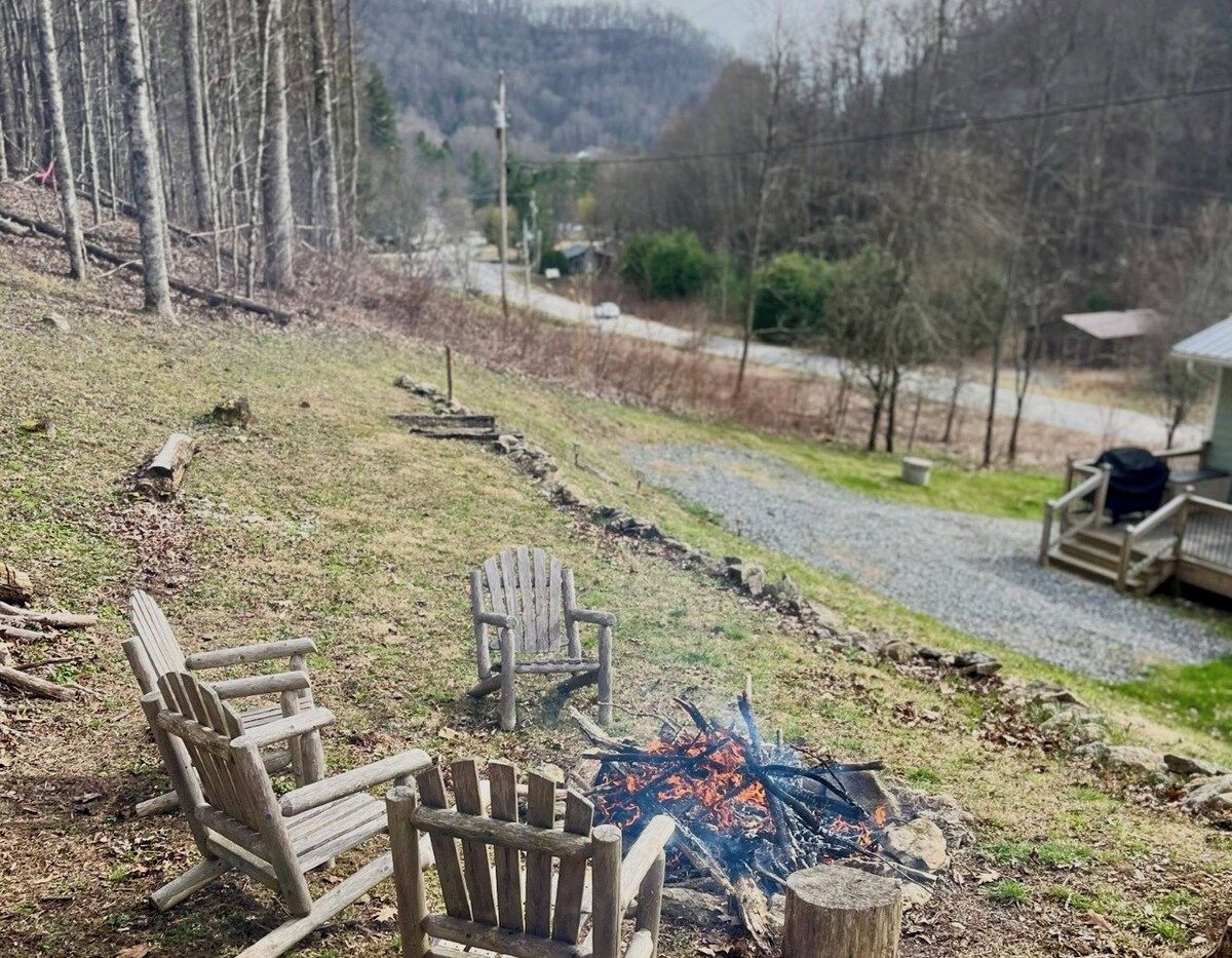 A fire pit with two Adirondack chairs in a grassy outdoor area with a view of a road and buildings in the distance.
