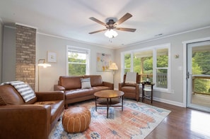 A living room with a brown leather sofa, a round coffee table, and a patterned rug. There is a ceiling fan, a fireplace, and a view through large windows. - Mountain Getaway | Fire Pit, Mtn Scenery, Stars (Mars Hill)