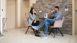 Un interior moderno con dos personas sentadas en sillas, una mesa con un objeto decorativo y una pared con un diseño geométrico.