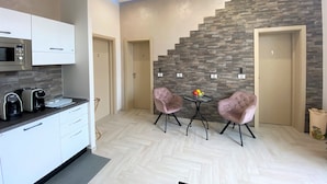 Cocina moderna con armarios blancos, un microondas integrado y una cafetera. El suelo presenta un patrón de espiga y una escalera con una pared de piedra. Dos sillas rosas están colocadas en una pequeña mesa con fruta.