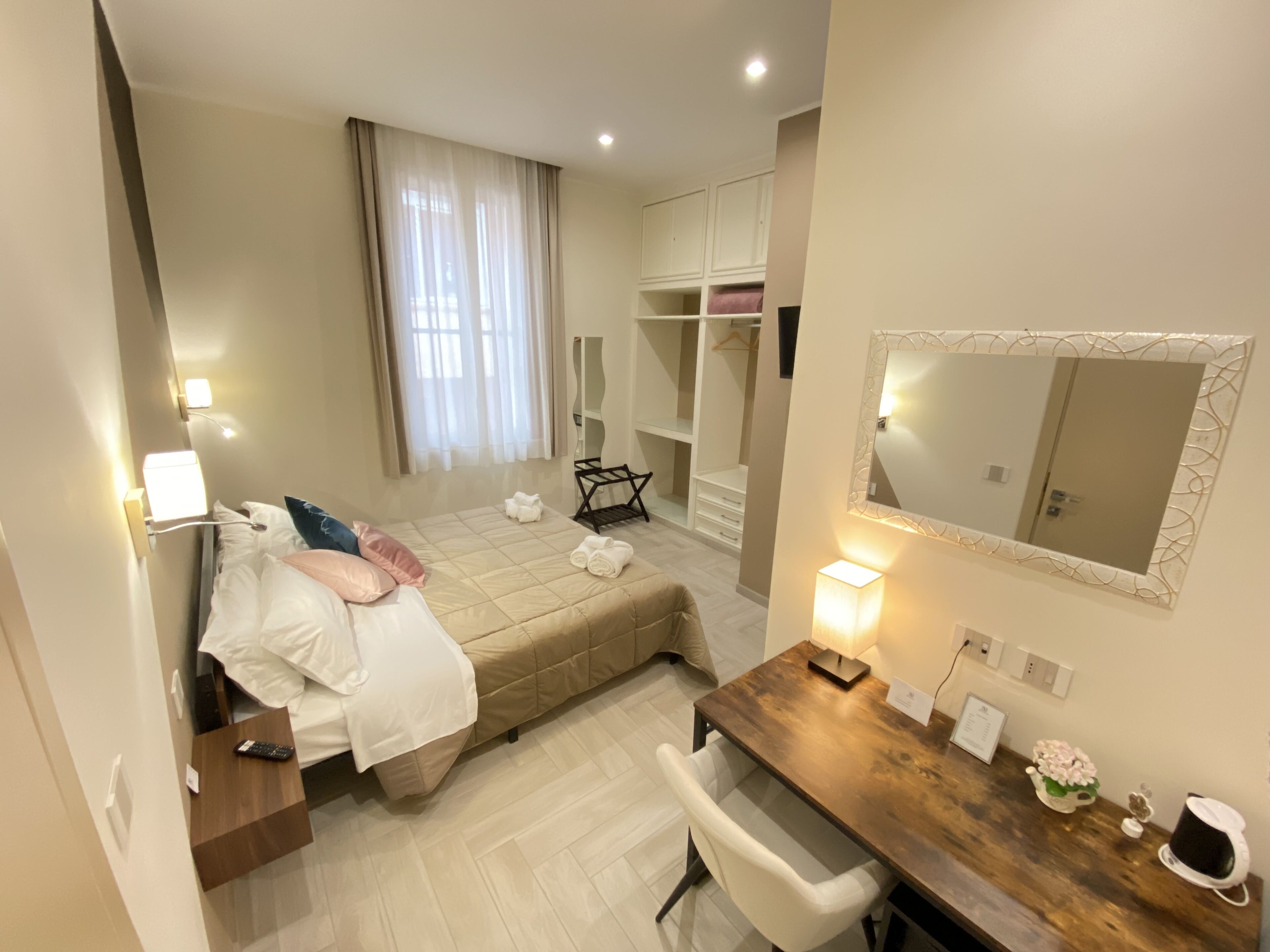 Habitación de hotel con cama, mesita de noche, silla, escritorio, espejo, armario y ventana con cortinas.