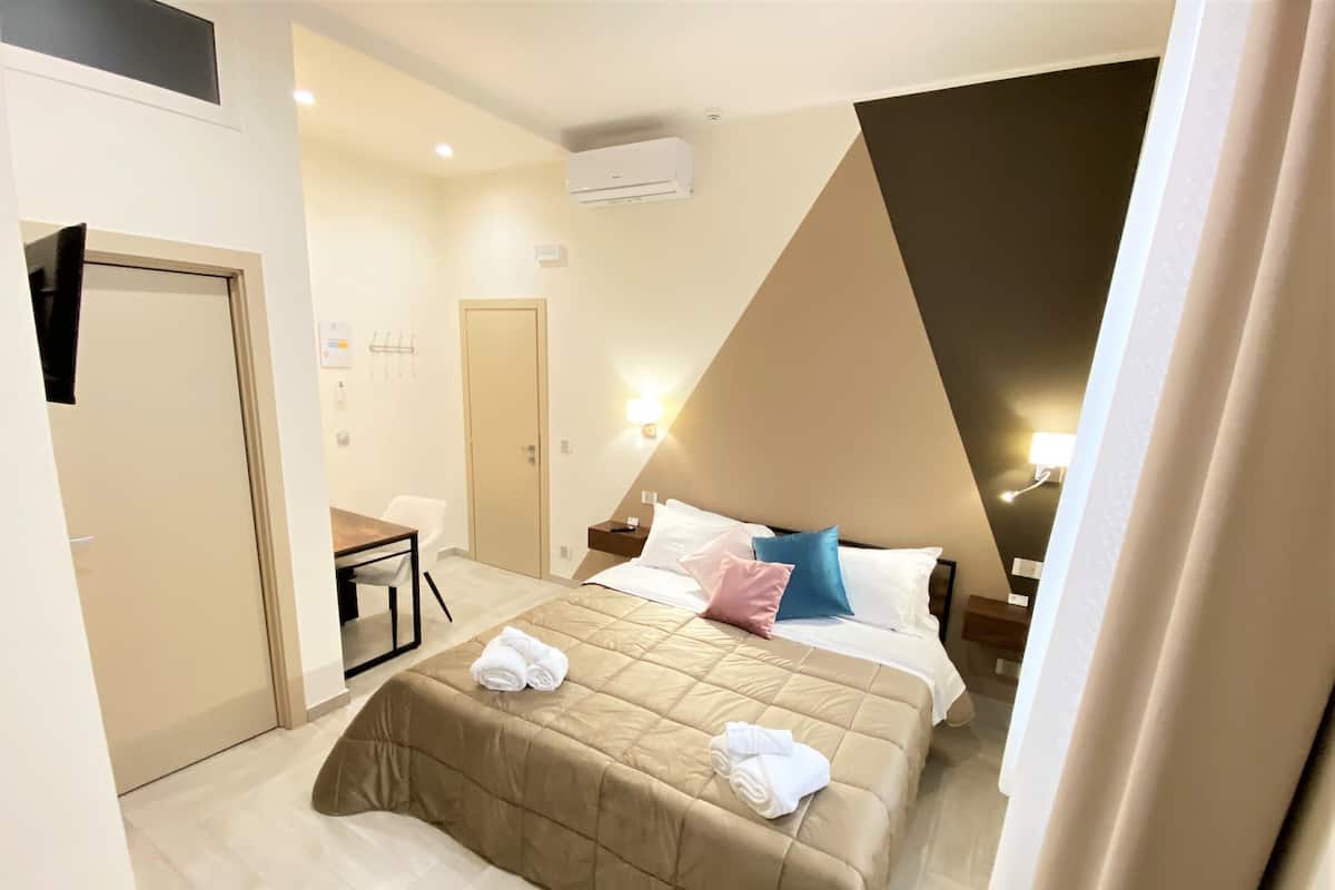 A hotel room with a bed, a desk, a chair, a door, and an air conditioning unit.