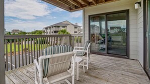 A covered outdoor deck with two white wicker chairs, a glass door leading to a covered area, and a view of neighboring houses. - Cordgrass Bay 2315-E (Wrightsville Beach)