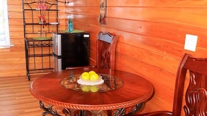 A wooden interior with a round dining table, chairs, a small refrigerator, and a wall-mounted light fixture.