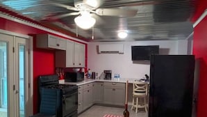 Eine kompakte Küche mit Gasherd, Mikrowelle und Kühlschrank. Ein Esstisch mit kariertem Tischläufer und ein Stuhl stehen bereit. Der Raum verfügt über eine rote Akzentwand und einen Deckenventilator.