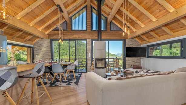 Ein großzügiger Wohnbereich mit hoher Decke, Holzbalken, Kamin, einer Couch und einem Essbereich mit Stühlen und einem Tisch.